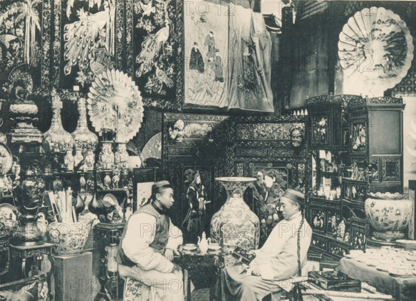 View of a Chinese shop at the Antwerp World Exhibition in 1894, Magasin Chinois, Exposition Universelle d'Anvers 1894, page, photomechanical print, photographer: Charles Bernhoeft, Antwerp, cliché maker: Charles Bernhoeft, Luxembourg (Belgian province), 1894, Charles Bernhoeft, photographer, cliché maker, paper, collotype, photoprint, height 153 mm x width 216 mm, Print on third unnumbered page after page 46., shop, store, world fair, world exhibition, World Exhibition 1894 (Antwerp), Period, 1894-05-05 - 1894-11-05, A crowded interior scene filled with decorative objects and furniture, including large ornate vases, carved wooden cabinets with intricate panels, folding screens, and patterned textiles hanging on the walls. Two men wearing traditional clothing and hats sit facing each other at a small table with a teapot and cups, one holding a small box while the other leans forward. Decorative fans and parasols with painted designs are displayed on the walls, and shelves and stands hol...