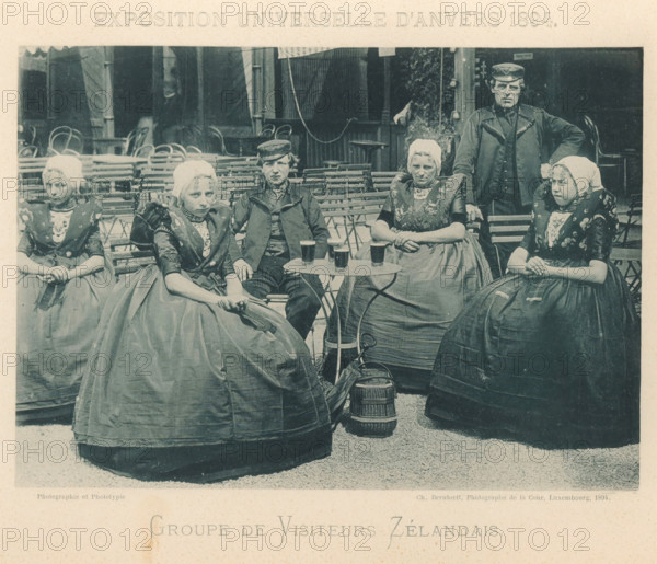 Group of visitors from Zeeland at the Antwerp World Exhibition in 1894, Groupe de Visiteurs Zélandais, Exposition Universelle d'Anvers 1894, page, photomechanical print, photographer: Charles Bernhoeft, Antwerp, cliché maker: Charles Bernhoeft, Luxembourg (Belgian province), 1894, Charles Bernhoeft, photographer, cliché maker, paper, collotype, photoprint, height 154 mm x width 217 mm, anonymous historical persons depicted in a group, in a group-portrait, world fair, world exhibition, World Exhibition 1894 (Antwerp), Period, 1894-05-05 - 1894-11-05, Five women and one man sit and stand around folding chairs and small round tables in an outdoor setting. The women wear full skirts, patterned bodices, and white caps, with layered collars and decorative embroidery on their garments. The man wears a dark jacket and a cap and stands with one hand resting on a chair. On the table are several glasses filled with a dark beverage. Behind them are stacked and arranged folding chairs and parts of ...