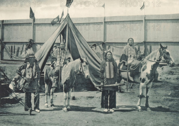 Members of Pawnee Bill's group at the Antwerp World Exhibition in 1894, Types de la Troupe de Pawne Bill, Chefs Jouws avec leurs femmes, Exposition Universelle d'Anvers 1894, page, photomechanical print, photographer: Charles Bernhoeft, Antwerp, Luxembourg (Belgian province), 1894, paper, collotype, photoprint, height 153 mm x width 216 mm, Print on third unnumbered page after page 62., horse, indigenous peoples of North America, world fair, world exhibition, World Exhibition 1894 (Antwerp), Period, 1894-05-05 - 1894-11-05, A group of people posed in front of a tent with a tall triangular entrance and a wooden fence behind it; two horses stand near the tent, one light-colored and one with darker patches, both wearing bridles and saddles; several adults wear patterned clothing and long braided hair, one person standing in front holding a cylindrical object, another wearing a feathered headdress, and one seated on a horse holding a child wrapped in cloth; flags fly above the fence and fa...
