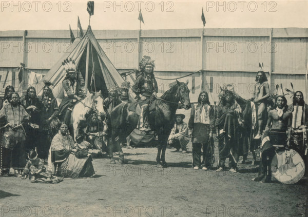 Types Guérriers de la Troupe de Pawnee Bill, Members of Pawnee Bill's group at the Antwerp World Exhibition in 1894, Exposition Universelle d'Anvers 1894, page, photomechanical print, photographer: Charles Bernhoeft, Antwerp, Luxembourg (Belgian province), 1894, paper, collotype, photoprint, height 152 mm x width 219 mm, horse, indigenous peoples of North America, world fair, world exhibition, World Exhibition 1894 (Antwerp), Period, 1894-05-05 - 1894-11-05, A wide group of people arranged in front of a large wooden fence and a canvas tipi, many wearing feathered headdresses and traditional garments, with several individuals mounted on horses and others standing or seated on the ground; visible accessories include beaded necklaces, fringed clothing, patterned blankets, and a round shield resting near one person, while small flags fly atop poles behind the tipi and a variety of long hair styles and face paint appear among the group.