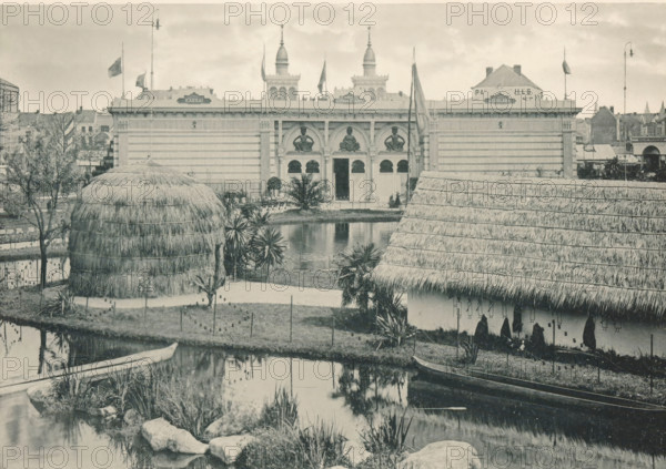 View of a recreated Congolese village at the Antwerp World Exhibition in 1894, Palais du Congo, Exposition Universelle d'Anvers 1894, page, photomechanical print, photographer: Charles Bernhoeft, Antwerp, cliché maker: Charles Bernhoeft, Luxembourg (Belgian province), 1894, Charles Bernhoeft, photographer, cliché maker, paper, collotype, photoprint, height 151 mm x width 216 mm, Print on third unnumbered page after page 66., prospect of village, silhouette of village, civic architecture, edifices, dwellings, world fair, world exhibition, World Exhibition 1894 (Antwerp), Period, 1894-05-05 - 1894-11-05, A wide scene shows a body of water in the foreground with grassy banks and rocks, a small wooden canoe moored at the edge, and reflections of surrounding elements on the water surface; behind the water there are clustered plants and short palms, a large rounded structure made of layered thatch, and a long thatched roof building with visible eaves; further back stands a formal striped bui...