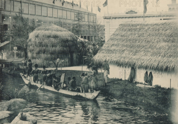 Congo, Vue du Village, Men in a rowing boat in a recreated Congolese village at the Antwerp World Exhibition in 1894, Exposition Universelle d'Anvers 1894, page, photomechanical print, photographer: Charles Bernhoeft, Antwerp, cliché maker: Charles Bernhoeft, Luxembourg (Belgian province), 1894, Charles Bernhoeft, photographer, cliché maker, paper, collotype, photoprint, height 154 mm x width 216 mm, adult man, prospect of village, silhouette of village, world fair, world exhibition, rowing-boat, canoe, etc. (four persons or more, traffic and transport), World Exhibition 1894 (Antwerp), Period, 1894-05-05 - 1894-11-05, A waterside scene shows a long narrow boat filled with people wearing minimal clothing and holding paddles, positioned near the grassy bank; behind them are two large thatched structures with thick straw roofs, one rounded and dome-like and the other with a steep sloping roof, flanked by tropical plants and small trees; a man in lighter clothing and a hat stands near the...