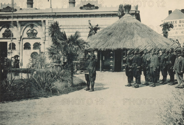 Congo, Troupe indigène, Group of men with instruments and weapons in a recreated Congolese village at the Antwerp World Exhibition in 1894, Exposition Universelle d'Anvers 1894, page, photomechanical print, photographer: Charles Bernhoeft, Antwerp, cliché maker: Charles Bernhoeft, Luxembourg (Belgian province), 1894, Charles Bernhoeft, photographer, cliché maker, paper, collotype, photoprint, height 154 mm x width 216 mm, Print on third unnumbered page after page 70., adult man, firearms, prospect of village, silhouette of village, horn, trumpet, cornet, trombone, tuba, world fair, world exhibition, World Exhibition 1894 (Antwerp), Period, 1894-05-05 - 1894-11-05, A sepia-toned scene shows a group of uniformed men standing in formation holding long rifles with bayonets, lined up beside a large round thatched hut with a conical roof; two younger boys stand in front holding a pair of curved objects. In the left background there is a pale ornate building facade with arched openings and sc...