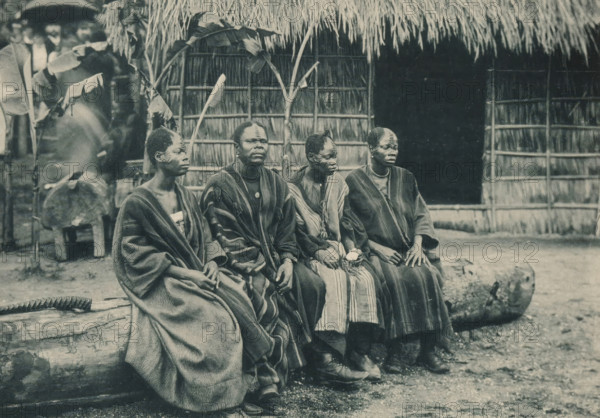 Groupe de Congolais, Four men sit on a tree trunk in a recreated Congolese village at the Antwerp World Exhibition in 1894, Exposition Universelle d'Anvers 1894, page, photomechanical print, photographer: Charles Bernhoeft, Antwerp, cliché maker: Charles Bernhoeft, Luxembourg (Belgian province), 1894, Charles Bernhoeft, photographer, cliché maker, paper, collotype, photoprint, height 153 mm x width 216 mm, adult man, civic architecture, edifices, dwellings, world fair, world exhibition, beam, log of wood, World Exhibition 1894 (Antwerp), Period, 1894-05-05 - 1894-11-05, Four seated people wearing long patterned garments sit side by side on a carved log in front of a thatched structure with woven walls, with broad leaves and plant stalks visible behind them; their faces show decorative scarification, and one person holds a small wrapped bundle while another wears a round pendant; a blurred figure and a drum appear in the background to the left and a length of rope lies on the log near t...