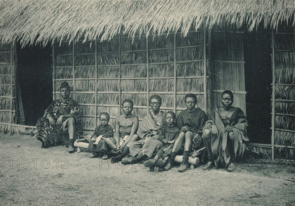 A family sits in front of a house in a recreated Congolese village at the Antwerp World Exhibition in 1894, Famille Congolaise, Exposition Universelle d'Anvers 1894, page, photomechanical print, photographer: Charles Bernhoeft, Antwerp, cliché maker: Charles Bernhoeft, Luxembourg (Belgian province), 1894, Charles Bernhoeft, photographer, cliché maker, paper, collotype, photoprint, height 153 mm x width 216 mm, Print on third unnumbered page after page 74., civic architecture, edifices, dwellings, children (first degree family relationships), world fair, world exhibition, parents (first degree family relationships), World Exhibition 1894 (Antwerp), Period, 1894-05-05 - 1894-11-05, A group of people sit in a row on a low wooden bench in front of a woven thatch wall and a deep overhanging thatch roof, with two dark doorways visible behind them; the thatch panels show vertical and horizontal supports and bundled reeds. The seated figures wear a mix of loose garments and patterned cloth, so...