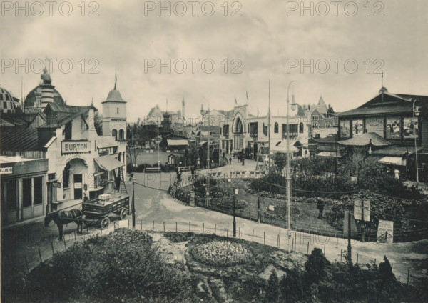 Quartier Oriental, View of the Oriental part of the Antwerp World Exhibition in 1894, Exposition Universelle d'Anvers 1894, page, photomechanical print, photographer: Charles Bernhoeft, Antwerp, cliché maker: Charles Bernhoeft, Luxembourg (Belgian province), 1894, Charles Bernhoeft, photographer, cliché maker, paper, collotype, photoprint, height 153 mm x width 216 mm, Print on third unnumbered page after page 78., prospect of village, silhouette of village, world fair, world exhibition, World Exhibition 1894 (Antwerp), Period, 1894-05-05 - 1894-11-05, A wide black-and-white street scene shows a curved road flanked by a low fenced garden in the foreground and a row of buildings and pavilions in the background. On the left, a horse-drawn wagon is parked near storefronts with awnings and a small tower-like structure rising behind them; domed and striped roof shapes are visible further back. At center, a plaza area contains lamp posts, benches, trees, and an arched entrance on a larger bu...