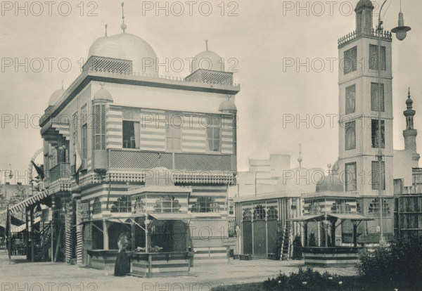Seccion Marocaine, View of the Moroccan part of the Antwerp World Exhibition in 1894, Exposition Universelle d'Anvers 1894, page, photomechanical print, photographer: Charles Bernhoeft, Antwerp, cliché maker: Charles Bernhoeft, Luxembourg (Belgian province), 1894, Charles Bernhoeft, photographer, cliché maker, paper, collotype, photoprint, height 154 mm x width 222 mm, counter, shop, prospect of village, silhouette of village, world fair, world exhibition, World Exhibition 1894 (Antwerp), Period, 1894-05-05 - 1894-11-05, A sepia-toned street scene showing a multi-story building with striped exterior bands, ornate latticework, rounded domes on the roof, and decorative finials, adjacent to a tall tower with patterned panels and a lamp fixture at its top; the foreground includes a small striped kiosk or stall with a person standing beside it, an awninged walkway with stairs, various windows and balcony railings, a gated entrance with arched openings, and low planted greenery along the fro...