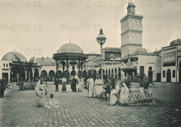 Tunisie, Une place a Tunis, View of a square in the Tunisian part of the Antwerp World Exhibition in 1894, Exposition Universelle d'Anvers 1894, page, photomechanical print, photographer: Charles Bernhoeft, Antwerp, cliché maker: Charles Bernhoeft, Luxembourg (Belgian province), 1894, Charles Bernhoeft, photographer, cliché maker, paper, collotype, photoprint, height 153 mm x width 216 mm, Print on third unnumbered page after page 82., prospect of city, town panorama, silhouette of city, square, circus, world fair, world exhibition, World Exhibition 1894 (Antwerp), Period, 1894-05-05 - 1894-11-05, A wide cobblestone courtyard with people wearing long draped garments gathered around a central tiled fountain and seated on its edge, framed by potted plants; a domed pavilion with ornate arches and decorative roof finials stands to the left of a tall multi-tiered tower with carved facade details, and a large decorative lamp post rises near the pavilion; arcaded colonnades, archways, and low...