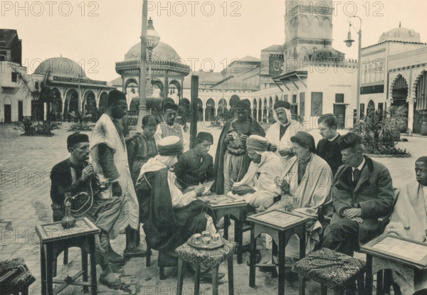 Tunisia, Partie de Cartes devant un Café arabe, Group of men playing a card game on a square in the Tunisian part of the Antwerp World Exhibition in 1894, Exposition Universelle d'Anvers 1894, page, photomechanical print, photographer: Charles Bernhoeft, Antwerp, cliché maker: Charles Bernhoeft, Luxembourg (Belgian province), 1894, Charles Bernhoeft, photographer, cliché maker, paper, collotype, photoprint, height 152 mm x width 217 mm, adult man, card games, square, circus, world fair, world exhibition, World Exhibition 1894 (Antwerp), Period, 1894-05-05 - 1894-11-05, A group of people sit and stand in an open courtyard surrounded by arched buildings and domed structures, with a tall ornate lamp post and a fountain visible in the background; many of the individuals wear headwraps, cloaks, robes, and traditional hats, while one person holds a long curved pipe connected to a vessel on a small table and several others hold small cups; there are low wooden tables and stools with patterned...