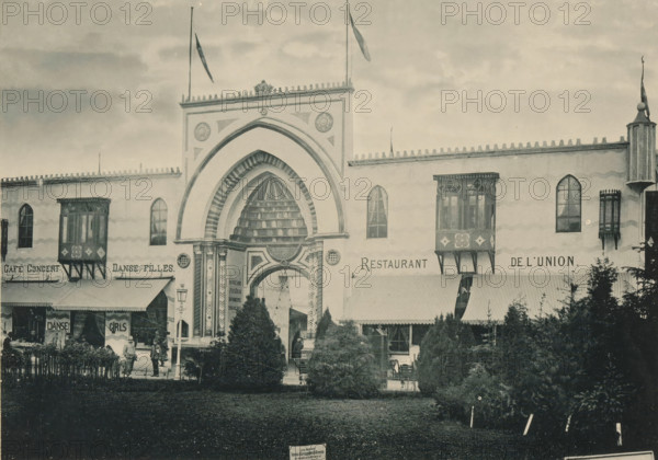 Palais Syrien, View of a Syrian palace, recreated at the Antwerp World Exhibition in 1894, Exposition Universelle d'Anvers 1894, page, photomechanical print, photographer: Charles Bernhoeft, Antwerp, cliché maker: Charles Bernhoeft, Luxembourg (Belgian province), 1894, Charles Bernhoeft, photographer, cliché maker, paper, collotype, photoprint, height 153 mm x width 215 mm, Print on third unnumbered page after page 86., palace, world fair, world exhibition, World Exhibition 1894 (Antwerp), Period, 1894-05-05 - 1894-11-05, A large building facade with a central ornate arched entrance featuring layered arch detailing and decorative columns, flanked by two projecting enclosed balconies with patterned panels and supporting brackets, several tall narrow arched windows, flagpoles with flags on the roofline, a row of decorative crenellations along the top edge, striped awnings over ground-level openings, a foreground area with trimmed shrubs and trees, a few people standing near the entrance ...
