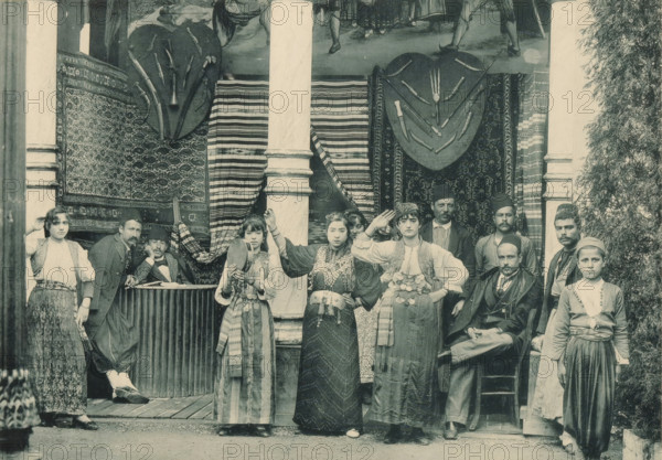 Syrie, Groupe de Danseuses, Group of dancers in the Syrian part of the Antwerp World Exhibition in 1894, Exposition Universelle d'Anvers 1894, page, photomechanical print, photographer: Charles Bernhoeft, Antwerp, cliché maker: Charles Bernhoeft, Luxembourg (Belgian province), 1894, Charles Bernhoeft, photographer, cliché maker, paper, collotype, photoprint, height 153 mm x width 216 mm, female dancer, male dancer, world fair, world exhibition, World Exhibition 1894 (Antwerp), Period, 1894-05-05 - 1894-11-05, A group of people posed in front of a doorway framed by two columns and patterned hanging textiles, with ornamental shields and curved objects mounted above. Several women in traditional layered garments and decorative belts stand at the front, two holding hand drums and one raising a hand to her head. Men around them wear a mix of suits, jackets, and traditional caps, with some seated and others standing beside or behind the women. A man leans on a counter partially covered by cl...