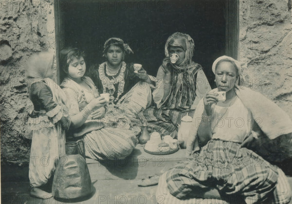 Four women drinking coffee in the Tunisian part of the Antwerp World Exhibition in 1894, Tunisia, Femmes prenant le Café, Exposition Universelle d'Anvers 1894, page, photomechanical print, photographer: Charles Bernhoeft, Antwerp, cliché maker: Charles Bernhoeft, Luxembourg (Belgian province), 1894, Charles Bernhoeft, photographer, cliché maker, paper, collotype, photoprint, height 153 mm x width 216 mm, Print on third unnumbered page after page 90., adult woman, child, cup of coffee, world fair, world exhibition, World Exhibition 1894 (Antwerp), Period, 1894-05-05 - 1894-11-05, Five seated people and a standing child occupy a dark doorway surrounded by rough stone walls, each holding small decorated cups. The figures wear layered, patterned clothing and head coverings in various styles, with jewelry visible on some. A tray with a teapot, lidded container, and stacked cups sits on the ledge in front of them, alongside additional vessels and a large carved head resting on the ground. Th...