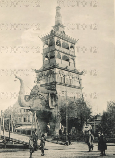 View of an elephant with a building on its back, recreated at the Antwerp World Exhibition in 1894, L'Eléphant Géant, Exposition Universelle d'Anvers 1894, page, photomechanical print, photographer: Charles Bernhoeft, Antwerp, Luxembourg (Belgian province), 1894, paper, collotype, photoprint, height 214 mm x width 153 mm, Print on third unnumbered page after page 94., trunked animals: elephant, civic architecture, edifices, dwellings, world fair, world exhibition, World Exhibition 1894 (Antwerp), Period, 1894-05-05 - 1894-11-05, A large ornate multi-tiered tower rises above a street scene, its tiers decorated with arched openings, carved details, and a bottle-shaped finial at the top; in front of the tower stands a giant elephant figure with a raised trunk and visible tusks, an ornamental howdah on its back, and a small statue mounted atop the howdah; scaffolding and ramps surround the elephant base, while groups of people in period clothing stand and walk nearby on a paved area, some ...