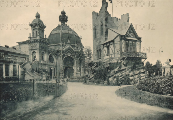 Moselburg, View of a house at the Antwerp World Exhibition in 1894, Exposition Universelle d'Anvers 1894, page, photomechanical print, photographer: Charles Bernhoeft, Antwerp, cliché maker: Charles Bernhoeft, Luxembourg (Belgian province), 1894, Charles Bernhoeft, photographer, cliché maker, paper, collotype, photoprint, height 153 mm x width 216 mm, civic architecture, edifices, dwellings, world fair, world exhibition, World Exhibition 1894 (Antwerp), Period, 1894-05-05 - 1894-11-05, A wide view of a landscaped area with a grand ornate building on the left featuring a large domed roof, a decorative tower, arched entrances, elaborate sculptural details, and a broad staircase leading down to a paved path; to the right of that building a half-timbered structure sits atop jagged rock-like forms adjacent to a tall ruined stone tower, with balustraded balconies and latticed windows visible; the foreground shows a curving paved walkway flanked by low posts and planted beds with shrubs and f...
