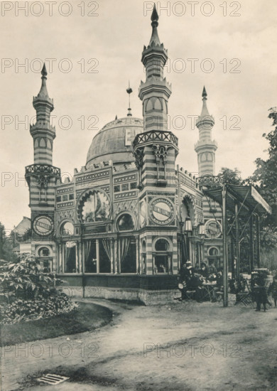 View of the pavilion of E. de Beukelaer & Cie at the Antwerp World Exhibition in 1894, Pavillon E. De Beukelaer et Cie, Exposition Universelle d'Anvers 1894, page, photomechanical print, photographer: Charles Bernhoeft, Antwerp, cliché maker: Charles Bernhoeft, Luxembourg (Belgian province), 1894, Charles Bernhoeft, photographer, cliché maker, paper, collotype, photoprint, height 214 mm x width 152 mm, palace, world fair, world exhibition, World Exhibition 1894 (Antwerp), Period, 1894-05-05 - 1894-11-05, A striped building with ornate arched windows and decorative panels topped by a large dome and several tall pointed minarets, each with layered segments and finials, stands beside a curved path and a planted area with low vegetation; the façade shows circular and scalloped window designs and columns with patterned capitals, and a covered outdoor seating area with chairs and tables where a small group of people sits, while trees frame the scene on the right.