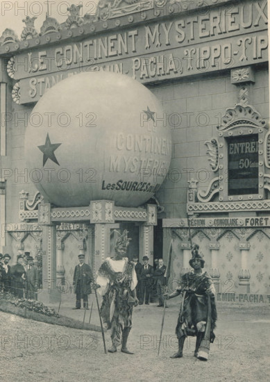 Two men in front of a diorama at the Antwerp World Exhibition in 1894, Le Continent mistérieux, Diorama, Exposition Universelle d'Anvers 1894, page, photomechanical print, photographer: Charles Bernhoeft, Antwerp, cliché maker: Charles Bernhoeft, Luxembourg (Belgian province), 1894, Charles Bernhoeft, photographer, cliché maker, paper, collotype, photoprint, height 215 mm x width 152 mm, adult man, world fair, world exhibition, diorama, World Exhibition 1894 (Antwerp), Period, 1894-05-05 - 1894-11-05, A large spherical object mounted on an ornate platform dominates the upper left, decorated with star shapes and sitting against a façade with carved ornamental trim and patterned panels; two figures in the foreground wear fur and feathered headdresses and hold long staffs while standing on a gravel surface, flanked by formally dressed men and uniformed attendants near the entrance of the decorated building, which features carved columns, scrollwork, and framed openings.