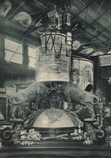 Monument to the Liebig Extract of Meat Company at the Antwerp World Exhibition in 1894, Monument de la Compagnie Liebig, Londres, Exposition Universelle d'Anvers 1894, page, photomechanical print, photographer: Charles Bernhoeft, Antwerp, cliché maker: Charles Bernhoeft, Luxembourg (Belgian province), 1894, Charles Bernhoeft, photographer, cliché maker, paper, collotype, photoprint, height 215 mm x width 152 mm, monument, statue, bull, world fair, world exhibition, World Exhibition 1894 (Antwerp), Period, 1894-05-05 - 1894-11-05, A monochrome indoor scene showing a large decorative display with a domed globe at its base and two life-sized bull sculptures positioned on either side, heads lowered toward the globe. Above the globe a tall cylindrical column rises with ornate embellishments and a bust perched near the top, surrounded by medallions hanging from ribbons. The ceiling overhead has a grid of beams and circular painted panels, and tall windows with vertical panes let light into t...