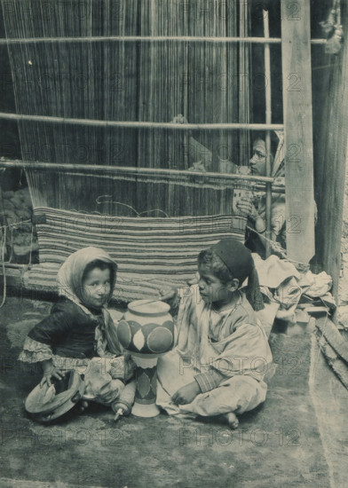 Tunisia, Carpet Manufacturer, Two children playing in front of a woman behind a loom in the Tunisian section of the Antwerp World Exhibition in 1894, Exposition Universelle d'Anvers 1894, page, photomechanical print, photographer: Charles Bernhoeft, Antwerp, clichémaker: Charles Bernhoeft, Luxembourg (Belgian province), 1894, Charles Bernhoeft, photographer, clichémaker, paper, collotype, photogravure, height 215 mm x width 153 mm, tambourine, child, drum (musical instrument), world fair, world exhibition, weaving-loom (women (labourers)), World Exhibition 1894 (Antwerp), Period, 1894-05-05 - 1894-11-05, A woven vertical warp and horizontal rods fill the upper background, with a striped woven textile hanging across the middle; a child peeks through the warp at the right, hand grasping a rod, headscarf visible; in the foreground two children sit on the ground beside a tall painted wooden object, one child wearing a headscarf and layered clothing holding a shallow bowl and a tambourine-l...