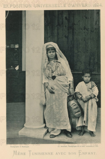 Mother and son in front of a building in the Tunisian part of the Antwerp World Exhibition in 1894, Mère Tunisienne avec son Enfant, Exposition Universelle d'Anvers 1894, page, photomechanical print, photographer: Charles Bernhoeft, Antwerp, cliché maker: Charles Bernhoeft, Luxembourg (Belgian province), 1894, Charles Bernhoeft, photographer, cliché maker, paper, collotype, photoprint, height 215 mm x width 152 mm, Print on third unnumbered page after page 138., boy (child between toddler and youth), mother with son(s) and daughter(s) (mother-love), world fair, world exhibition, World Exhibition 1894 (Antwerp), Period, 1894-05-05 - 1894-11-05, A monochrome photograph shows a woman standing beside a large round column and a child standing next to her with a wooden door as the background; the woman wears a long draped garment and a head covering that falls over her shoulders, layered necklaces and bracelets, and holds a large jug at her side while her feet are shod in closed shoes, and t...