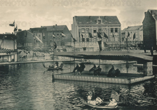 View of a floating platform with dancers at the Antwerp World Exhibition in 1894, Cirque naval du Captaine Boyton (Danse serpentine), Exposition Universelle d'Anvers 1894, page, photomechanical print, photographer: Charles Bernhoeft, Antwerp, cliché maker: Charles Bernhoeft, Luxembourg (Belgian province), 1894, Charles Bernhoeft, photographer, cliché maker, paper, collotype, photoprint, height 152 mm x width 216 mm, rowing-boat, canoe, group dancing, circus, world fair, world exhibition, World Exhibition 1894 (Antwerp), Period, 1894-05-05 - 1894-11-05, A waterfront scene shows a calm body of water in front of a pier with a striped canopy covering a platform where several seated figures are visible, a small rowboat with a standing oarsman and seated passengers floats nearby, and another small boat in the foreground carries seated figures and a visible flag at its stern; behind the pier a wooden fence runs along the shore with buildings of varying heights and architectural details rising...