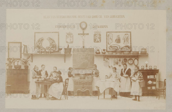 View of a home economics class, photograph, page, Antwerp, c. 1888 - in or before 1893, photographer, paper, daylight collodion silver print, height 106 mm x width 170 mm, cooking food, elementary school, class, form, household chores, A faded sepia photograph of an interior scene showing a wall with framed pictures, a clock, a crucifix, shelves with pots and pans, and hooks holding kitchen utensils; a central stove with a pot, a chalkboard above it, and a table covered with a cloth in front; a group of young girls and a woman wearing aprons standing and seated around tables and the stove, some holding bowls or utensils and some engaged with food on the tables; cabinets and sideboards on either side with jugs, dishes, and potted plants arranged on top; decorative plaques and framed meat diagrams mounted on the wall above the shelves.
