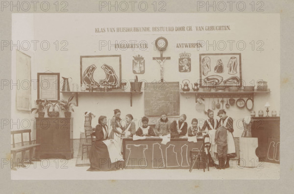 View of a home economics class, photograph, page, Antwerp, c. 1888 - in or before 1893, photographer, paper, daylight collodion silver print, height 106 mm x width 171 mm, elementary school, class, form, sewing, household chores, A sepia-toned classroom scene shows a group of girls and a woman seated and standing behind a long table covered with cloths and garment patterns, all wearing aprons and dresses in period style; shelves and a wall behind them display framed prints with stylized illustrations, kitchenware including pots, kettles, pitchers, pans, and a crucifix with a round clock above it, flanked by decorative crests, while plants in pots and framed items sit on a cabinet to the left and a small lamp and jars rest on a cabinet to the right, with chairs and a wire dress form visible at the edges.