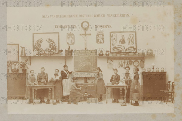 View of a home economics class, photograph, page, Antwerp, c. 1888 - in or before 1893, photographer, paper, daylight collodion silver print, height 105 mm x width 164 mm, elementary school, class, form, household chores, A sepia-toned indoor scene shows a row of young women and girls standing and working along a wall with shelves and framed illustrations, a crucifix, a wall clock, and decorative shields above them. The wall shelves hold metal pots, kettles, pans, and other kitchen utensils, and there are framed anatomical-style prints depicting cross-sections of meat. In front of the wall are tables, a stove, and a small blackboard propped on a mantel; various cooking tools and containers sit on the tables and stove. Several potted plants occupy a cabinet on the left, and jars and a lamp sit atop a cabinet on the right beside stacked chairs. The women wear long dresses and aprons; some hold utensils, a baking pan, a mop, or a bucket, while one kneels at the stove and another stands be...