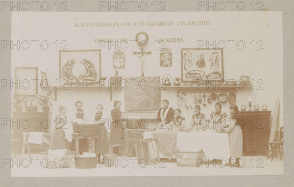 View of a home economics class, photograph, page, Antwerp, c. 1888 - in or before 1893, photographer, paper, daylight collodion silver print, height 104 mm x width 171 mm, elementary school, class, form, laundering, household chores, A faded sepia scene showing a room with a long shelf mounted on the wall holding framed pictures, framed diagrams of cuts of meat, pitchers, pots, kettles, and various kitchen utensils hanging beneath the shelf; a central wall clock above a crucifix and two heraldic shields; a chalkboard with handwritten entries mounted below the shelf; a group of girls standing and seated along the front of the room wearing aprons and dresses, some handling cloths and laundry items, with basins, wooden tubs, baskets, and a washboard on the floor; potted plants and a framed item on a side cabinet at one end and a lamp and jars on a cabinet at the other end, with a chair leaning against the wall.
