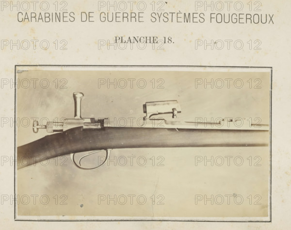 Barrel of a gun, photograph, page, number: 'Planche 18.', inscription: 'Carabines de guerres systèmes Fougeroux', photographer: Edouard Magis, Liège, c. 1857 - in or before 1867, paper, albumen print, height 96 mm x width 158 mm, Image in section at the back., firearms: rifle, A sepia-toned photograph framed by a thin dark border shows a close-up of a firearm mounted horizontally, featuring a dark wooden stock with a smooth curved surface, a metal trigger guard with an oval shape, a metal bolt mechanism with a vertical handle, a cylindrical knurled adjustment knob at the rear, and a rectangular sight mounted atop the barrel; the background is a lighter textured surface with small speckles and faint stains, and the paper surrounding the photograph shows age spots and worn edges.
