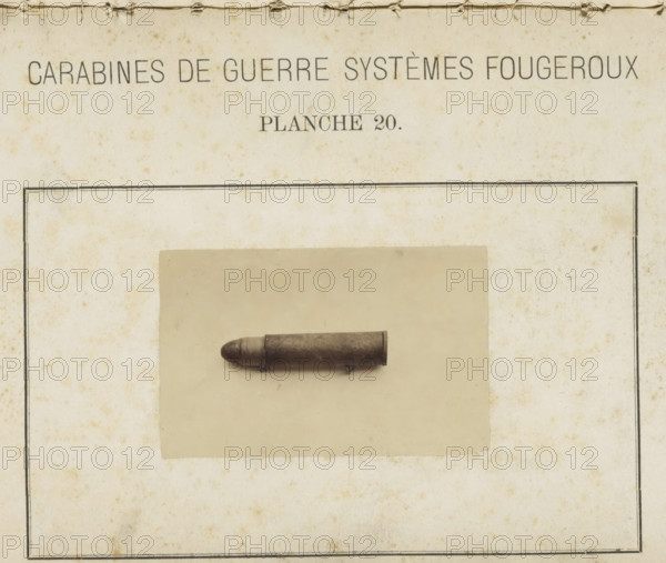 Bullet, photograph, page, number: 'Planche 20.', inscription: 'Carabines de guerres systèmes Fougeroux', photographer: Edouard Magis, Liège, c. 1857 - in or before 1867, paper, albumen print, height 55 mm x width 88 mm, Image in section at the back., ammunition, A yellowed rectangular page with a narrow dark border contains a centered faded photograph of a cylindrical metallic object with a rounded tip resting horizontally, mounted on a lighter rectangular matte; the page shows foxing and small brown spots, a torn top edge with visible fibers, and subtle creases and stains across its surface.