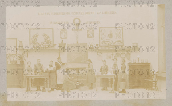 View of a home economics class, photograph, page, Antwerp, c. 1888 - in or before 1893, photographer, paper, daylight collodion silver print, height 104 mm x width 174 mm, elementary school, class, form, household chores, A faded sepia photograph of an interior room showing a long wall with framed decorative pictures, a clock, a crucifix, shelves holding kettles and pots, and hanging pans. Along the lower wall there are tables and a stove with a woman kneeling in front of it, another woman tending the stove, and several girls standing or working at tables holding utensils, bowls, a mop, and a bucket. Potted plants sit on a cabinet at one side and a lamp and jars sit on a tall piece of furniture at the other side, with chairs lined up beside it. The floor is visible and the room has a high window on the right.