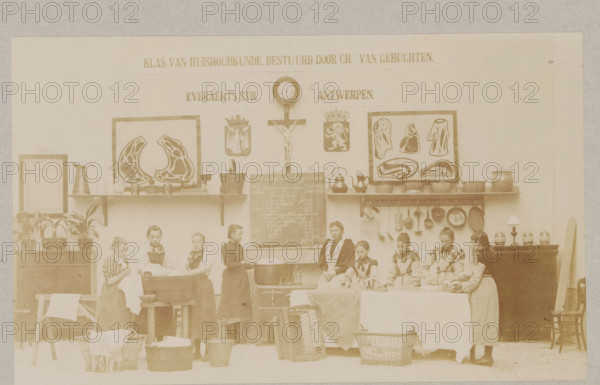 View of a home economics class, photograph, page, Antwerp, c. 1888 - in or before 1893, photographer, paper, daylight collodion silver print, height 103 mm x width 169 mm, elementary school, class, form, laundering, household chores, A sepia-toned indoor scene shows a group of young women and girls standing and working along a long table and near washbasins, wearing long dresses and aprons with some wearing dark pinafores and high collars, their hair pulled back. Above them shelves hold various pots, pitchers, kettles, pans, and decorative framed illustrations of food cuts and a crucifix with a round clock above it. Wall hooks display hanging utensils like ladles and strainers. On the floor and in front are wicker baskets, wooden tubs, and basins with cloths draped over them. Potted plants sit on a cabinet at one side and small jars and a lamp sit on a sideboard at the other side. A chalkboard with handwriting leans against the wall behind the group. The room has plain walls and a high...
