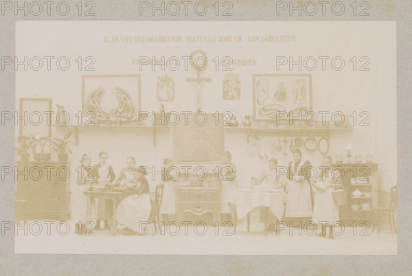 View of a home economics class, photograph, page, Antwerp, c. 1888 - in or before 1893, photographer, paper, daylight collodion silver print, height 104 mm x width 171 mm, cooking food, elementary school, class, form, household chores, A faded sepia photograph showing an interior scene with a long wall as a backdrop, shelves mounted on the wall holding pitchers, pots, kettles, bowls, and framed illustrations; a clock and a crucifix are mounted above a central stove with a chalkboard on top of it; potted plants sit on a cabinet at one side and jars and dishes sit on shelving at the other side; several girls and a woman wearing aprons stand and sit around tables and the stove, some holding bowls, utensils, or a basket, others appearing to be engaged in food preparation; chairs and tables with tablecloths are arranged in front of the stove and along the scene.