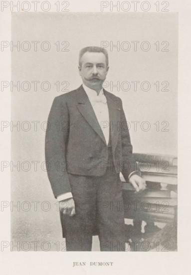 Portrait of Jean Dumont, Jean Dumont, page, photomechanical print, inscription: 'Similigravure.', cliché maker: Jean Malvaux, c. 1899 - in or before 1904, photographer, paper, autotypie, height 145 mm x width 101 mm, Person, historical persons, A sepia-toned portrait shows a man standing next to a small ornate table, wearing a dark suit jacket over a light waistcoat and a high-collared shirt with a bow tie, his hair neatly combed and a styled mustache visible, one hand resting on the table and the other hanging by his side, with a plain background behind him.