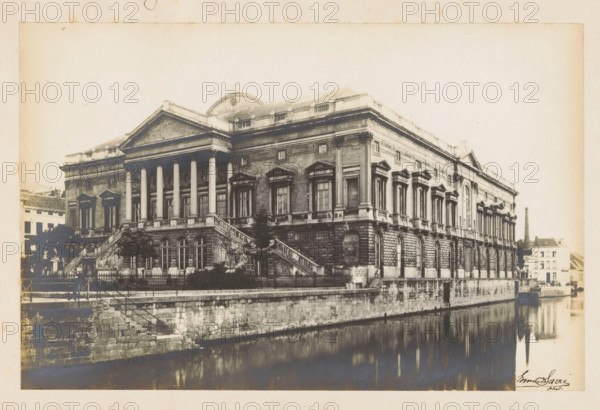 View of the courthouse in Ghent, photograph, page, inscription: 'Palais de Justice au Gand', photographer: Edmond Sacré, Ghent, c. 1894 - in or before 1899, paper, gelatin silver print, developing gelatin silver print, height 115 mm x width 177 mm, Photo on second unnumbered page after page 87., court building, Palace of Justice (1846-2007), A large classical stone building with a columned portico and triangular pediment sits beside a calm body of water, showing rows of tall rectangular windows with decorative pediments above them and a rusticated ground floor with arched openings, flanking exterior staircases leading up to the entrance, a balustraded terrace, ornamental cornices and pilasters along the façade, small trees and shrubs near the staircases, a low stone embankment at the water’s edge, a narrow canal reflecting the building, and a smaller multi-story structure and a tall chimney visible in the background.