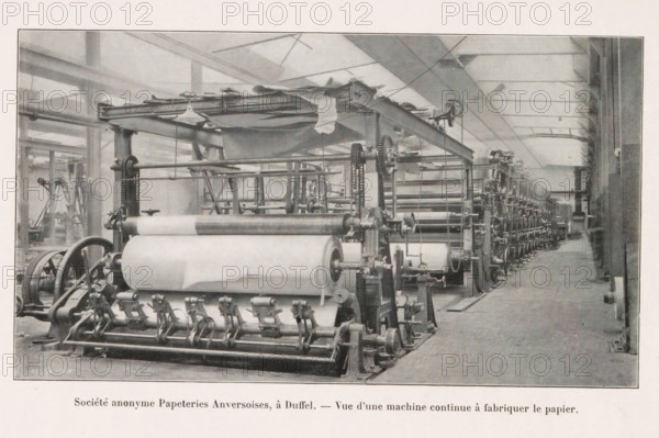 Société anonyme Papeteries Anversoises, à Duffel. - Vue d'une machine continu à fabriquer le papier, View of a paper-making machine at the Société anonyme Papeteries Anversoises in Duffel, page, photomechanical print, Duffel, cliché maker: Jean Malvaux, c. 1906 - in or before 1911, Jean Malvaux, photographer, cliché maker, paper, autotypie, height 100 mm x width 179 mm, Print on second unnumbered page after page 19., paper (writing material), machine driven by electricity, A wide industrial interior with a long row of large mechanical rollers and gears arranged along the floor, connected by metal frames and chains, with sheets of material wound around some rollers and draped fabric hanging above; the space has high ceilings with beams and skylights, tall vertical supports, and a clear aisle running alongside the machinery toward the far end; various crank handles, wheels, and pulleys are visible on the equipment, and the scene is rendered in a monochrome palette.
