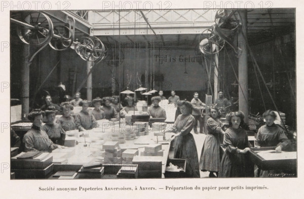 Société anonyme Papeteries Anversoises, à Duffel. - Vue d'une machine continuation à fabriquer le papier, Group of women preparing stacks of paper for small printing in the Société anonyme Papeteries Anversoises in Antwerp, page, photomechanical print, Antwerp, cliché maker: Jean Malvaux, c. 1906 - in or before 1911, Jean Malvaux, photographer, cliché maker, paper, autotypie, height 102 mm x width 172 mm, adult woman, paper (writing material), production of printed matter, book-production, A large indoor workspace with high ceilings and visible pulley wheels and belts near the ceiling, rows of long tables covered with stacks of rectangular sheets and boxes, and a group of women wearing long plain dresses standing and seated around the tables, some handling sheets and boxes and others looking toward the camera, with hanging lamps suspended above the central workspace and structural columns and beams visible in the background.