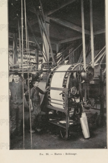 Two men and a paper bending machine in a printing shop in Haren, Belgium, page, photomechanical print, inscription: 'Fig. 16. - Haren: Bobinage.', monogram: 'AN', photographer: unknown, Haren (Brussels), cliché maker: Jean Malvaux, c. 1901 - in or before 1906, Brussels, paper, autotype, height 170 mm x width 110 mm, printing, printing office, adult man, workshop-interior, machine driven by electricity, A tall industrial interior with large beams and support columns fills the background, intersected by a network of ropes, pulleys, and metal frames. In the foreground a large cylindrical machine with a broad wrapped band is mounted on a metal stand with visible gears, wheels, and a drum; a long strip of material hangs from the machine to the floor. Two young boys stand beside the machinery, wearing work clothes and aprons; one boy faces forward holding a rope, the other stands closer to the machine with a hand resting on it, looking toward the first boy. The floor shows scattered tools an...