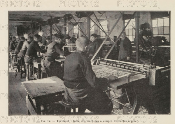 Men cut out printed playing cards in a printing house in Turnhout, page, photomechanical print, inscription: 'Fig. 17. - Turnhout: Salle des machines à couper les cartes à jouer.', monogram: 'AN', photographer: unknown, Turnhout, cliché maker: Jean Malvaux, c. 1901 - in or before 1906, paper, autotypie, height 110 mm x width 171 mm, printing, printing office, adult man, playing-cards, workshop-interior, A black and white photograph of an indoor workshop showing rows of people seated at long workbenches and stools, several facing away from the camera and one prominent worker in the foreground seated with his back toward the viewer and hands on a flat surface holding a patterned sheet, visible mechanical equipment and rollers on the right side of the image, overhead belts and pulleys running across the ceiling, tall windows along the back wall letting light into the room, structural columns and diagonal support beams between the workbenches, workers wearing dark clothing and aprons, shel...