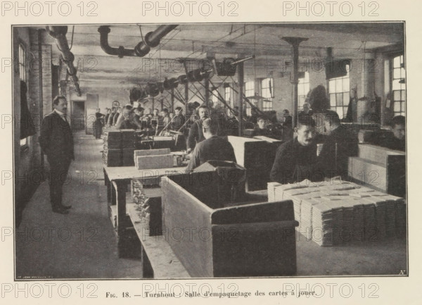 Men pack printed playing cards in a printing house in Turnhout, page, photomechanical print, monogram: 'AN', inscription: 'Fig. 18. - Turnhout: Salle d'empaquetage des cartes à jouer.', photographer: unknown, Turnhout, cliché maker: Jean Malvaux, c. 1901 - in or before 1906, unknown, Jean Malvaux, photographer, cliché maker, paper, autotypie, height 110 mm x width 170 mm, printing, printing office, adult man, playing-cards, workshop-interior, A wide interior scene shows a long room with rows of workbenches and stacks of rectangular bundles on and beside the benches. Men in dark clothing are seated and standing along the benches, some facing the camera and some turned toward the work, while a few figures stand in the aisle. Overhead pipes and a system of pulleys or belts run along the ceiling supported by columns, and light comes through tall multi-pane windows along the far wall. In the foreground there are large wooden partitions and neatly piled bundles on the benches, and the overal...