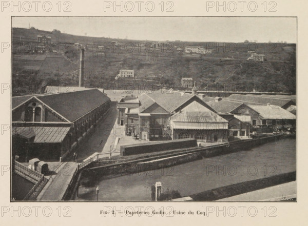 View of a building of the Godin paper factory in Huy, page, photomechanical print, inscription: 'Fig. 2. - Papeteries Godin: Usine du Coq', monogram: 'AN', photographer: unknown, Huy, cliché maker: Jean Malvaux, c. 1901 - in or before 1906, Hoei, paper, autotypie, height 110 mm x width 171 mm, factory-building, A sepia-toned aerial view of an industrial complex with long brick buildings and pitched roofs occupying the foreground, including a large structure with arched windows and a smaller building with a metal roof and decorative cupola, adjacent courtyards and paved walkways, a bridge leading into the site with a small vehicle and a lone pedestrian, a tall smokestack rising near the center, a stone wall separating the complex from an open water channel in front, and rolling hills in the background dotted with scattered houses and small farm buildings under a light sky.