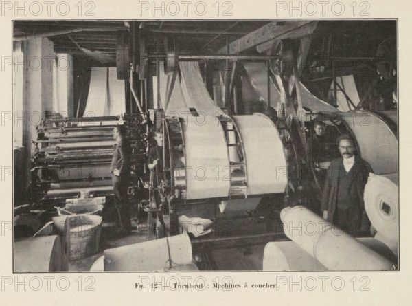 View of a workshop in a paper factory in Turnhout, page, photomechanical print, monogram: 'AN', inscription: 'Fig. 12. - Turnhout: Machines à coucher.', photographer: unknown, Turnhout, cliché maker: Jean Malvaux, c. 1901 - in or before 1906, paper, autotypie, height 110 mm x width 169 mm, workshop interior, A monochrome photograph shows an industrial interior with large rolling machines and wide rolls of material. Several vertical and horizontal shafts, gears, and rollers occupy the central area, with sheets of material running over and between them. Men stand among the machinery; one man is positioned near the foreground beside a large roll, another is partially visible behind a machine, and another stands further back by the rollers. Wooden beams and structural supports are visible overhead, and buckets or tubs sit on the floor near the machines. The image is framed by a light border and a lighter margin surrounds the photograph.