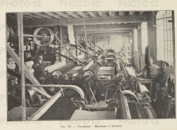 View of a workshop with brushing machines in a paper factory in Turnhout, page, photomechanical print, inscription: 'Fig. 13. - Turnhout: Machines à brosser.', monogram: 'AN', photographer: unknown, Turnhout, cliché maker: Jean Malvaux, c. 1901 - in or before 1906, paper, autotypie, height 110 mm x width 170 mm, workshop interior, machine driven by electricity, A wide interior view of a workshop filled with rows of large mechanical rollers and interconnected belts and pulleys arranged along the length of the room, with stacks of cylindrical rolls of material visible on shelves and along the sides, wooden ceiling beams overhead and vertical support posts, a long window letting light in from one side, and several workers standing near the machinery wearing work clothes and aprons.