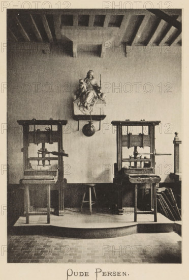 Old presses, Printing presses in the printing house of Christoffel Plantijn, page, photomechanical print, maker: Joseph Maes, c. 1881 - in or before 1886, paper, collotype, photoprint, height 160 mm x width 110 mm, Print on third unnumbered page after page 128., printing press, A monochrome interior photograph showing a wall with a mounted sculptural figure seated on a small ledge, holding objects and flanked by a round hanging ornament below it; beneath the sculpture are two tall wooden press-like structures with horizontal beams and mechanical components, separated by a wooden stool positioned between them; the floor in front has a raised platform with a curved front edge and visible floor tiles beyond it; the ceiling shows exposed wooden beams and a recessed rectangular pattern above the wall.