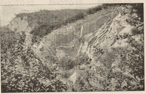 Carrière de Pierre-Pétru, montrant la torsion des couches, View of a quarry, page, photomechanical print, inscription: 'Reproduction direct du cliché photographique pris par MG Michelet, sous-lieutenant à l'École d'Application', maker: Michelet, M., c. 1887 - in or before 1892, paper, autotype, height 108 mm x width 175 mm, quarry, A grainy black-and-white photograph showing a steep eroded hillside with exposed tree roots and layered soil on the right, a sloping bank with a rough textured surface toward the center, and a foreground covered in scattered rocks and debris; vegetation appears along the top edge and mixed among the rocky foreground, and the image has a border with a lighter margin around it.