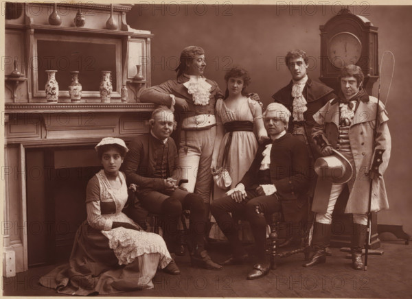 Group portrait of the members of The Vaudeville Company in 'The Road to Ruin', page, photomechanical print, photographer: Herbert Rose Barraud, c. 1881 - in or before 1886, paper, Woodburytype, height 114 mm x width 164 mm, Print two pages before page 178., Person, Kate Rorke, Charles Warner, James Fernandez, Thomas Thorne, Fuller Mellish, Fred Thorne, Louisa Peach, historical persons, adult woman, adult man, A group of seven people posed indoors in period clothing, arranged in two seated and five standing positions; the setting includes a mantelpiece with decorative vases and a large clock behind the group. The seated figures include a woman on the floor wearing a dark dress with a light apron and lace cap, and a man in dark formal attire with a wig and lace cuffs sitting on a carved wooden chair. Standing figures include a man in a light waistcoat and ruffled cravat leaning on the mantel, a young woman in a light gown with a dark sash standing beside him, a man in a dark coat with a ...