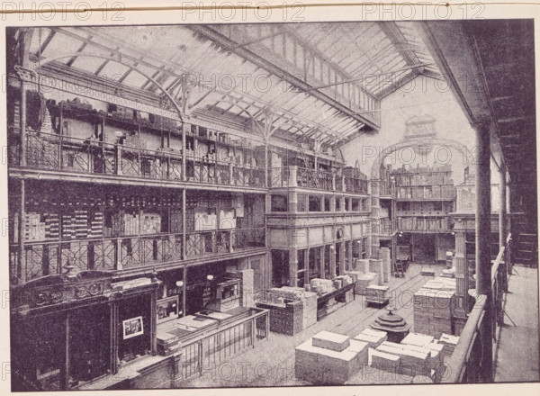 View of a Lutkie & Smit paper warehouse in Amsterdam, Lutkie & Smit Paper Warehouse in Amsterdam, page, photomechanical print, name: 'R...', cliché maker: unknown, c. 1887 - in or before 1892, photographer, paper, autotype, height 129 mm x width 187 mm, Print in a section of advertisements at the back of a book. Print is part of an advertising message by Lutkie & Smit., large-scale structure for storage, eg: warehouse, depot, silo, Amsterdam, A large interior space with a high arched roof supported by exposed beams and metal trusses, multiple tiers of open balconies with decorative railings lining the sides, tall columns rising between levels, stacked crates and boxes arranged on the floor, counters and shelving along the lower level, staircases and walkways connecting the balconies, hanging lamps and circular light fixtures suspended from the ceiling, architectural details such as ornate cornices and carved wood panels, and a curved archway visible at the far end.