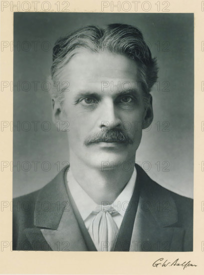 The right honorable Gerald William Balfour, photograph, page, inscription: 'Leeds', annotation, photographer: AC Hoskins, London, c. 1893 - c. 1903, paper, carbon print, height 228 mm x width 170 mm, Person, Gerald Balfour, historical persons (portraits and scenes from the life) ((full) bust portrait), A black and white portrait of a man from the shoulders up centered on a neutral gray background, showing a face with a neatly groomed mustache, high cheekbones, and deep-set eyes looking forward, with wavy hair swept back from a defined part at the top, visible ear on one side, and faint wrinkles on the forehead and around the eyes; he is wearing a dark jacket over a vest and a light-colored shirt with a tied neckcloth, and a light border surrounds the photograph.