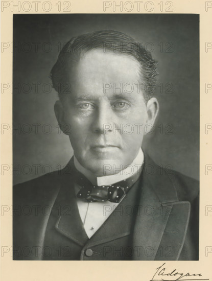The right honorable earl Cadogan KG, photograph, page, annotation, inscription: 'Dublin', photographer: Chancellor, Ireland, c. 1893 - c. 1903, paper, carbon print, height 228 mm x width 170 mm, Person, George Cadogan, historical persons (portraits and scenes from the life) ((full) bust portrait), A black and white portrait of an older man from the chest up centered in the frame, showing a close-up of his face with neatly combed hair, pronounced forehead, and neutral expression, his eyes looking forward and visible under subtle shadows, ears partially visible, and a smooth complexion with fine facial lines; he is wearing a dark suit jacket over a waistcoat and a white high collar, with a patterned bow tie at the throat; the photograph has a narrow light border and a darker background behind the man.