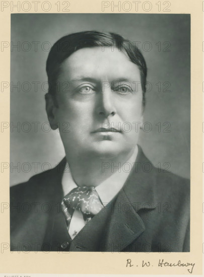 The right honorable Robert William Hanbury, photograph, page, annotation, inscription: '55, Baker St, London, W.', photographer: Elliott & Fry, c. 1893 - c. 1903, paper, carbon print, height 227 mm x width 171 mm, Person, Robert William Hanbury, historical persons (portraits and scenes from the life) ((full) bust portrait), A black and white portrait photograph of a man from the shoulders up, showing a close-up of his face with short, neatly combed hair, light-toned skin, and a calm expression. He is wearing a dark suit jacket over a vest and a white shirt, with a patterned necktie visible at the collar. The background is a plain, softly shaded backdrop, and the image has a narrow light-colored border around it.